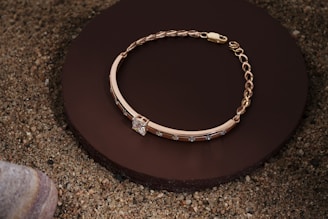 A gold bracelet with a chain and decorative stones is elegantly placed on a round, dark brown surface, surrounded by sand and a smooth stone in the corner.