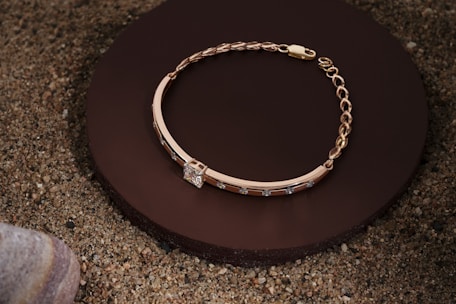 A gold bracelet with a chain and decorative stones is elegantly placed on a round, dark brown surface, surrounded by sand and a smooth stone in the corner.