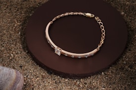 A gold bracelet with a chain and decorative stones is elegantly placed on a round, dark brown surface, surrounded by sand and a smooth stone in the corner.