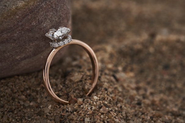 A delicate gold ring featuring a large diamond centerpiece surrounded by smaller diamonds is resting on coarse sand beside a smooth stone. The setting creates a contrast between the luxurious ring and the natural, earthy background.