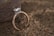A delicate gold ring featuring a large diamond centerpiece surrounded by smaller diamonds is resting on coarse sand beside a smooth stone. The setting creates a contrast between the luxurious ring and the natural, earthy background.