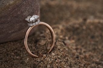 A delicate gold ring featuring a large diamond centerpiece surrounded by smaller diamonds is resting on coarse sand beside a smooth stone. The setting creates a contrast between the luxurious ring and the natural, earthy background.