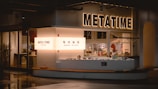 A modern coffee shop with the name 'Meta Time' prominently displayed in illuminated letters. The storefront has a sleek, minimalist design and features large windows that reveal a stylish interior with tables and potted plants. A counter is visible with coffee accessories and a menu.