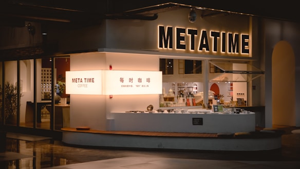 A modern coffee shop with the name 'Meta Time' prominently displayed in illuminated letters. The storefront has a sleek, minimalist design and features large windows that reveal a stylish interior with tables and potted plants. A counter is visible with coffee accessories and a menu.