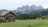 A rustic wooden cabin with a neatly stacked pile of firewood sits on a lush green meadow. In the background, there are majestic, jagged mountains with peaks shrouded in mist. Dense forests of evergreen trees stretch across the foothills. A few cows are grazing peacefully near the cabin, adding a pastoral touch to the serene landscape.