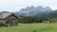 A rustic wooden cabin with a neatly stacked pile of firewood sits on a lush green meadow. In the background, there are majestic, jagged mountains with peaks shrouded in mist. Dense forests of evergreen trees stretch across the foothills. A few cows are grazing peacefully near the cabin, adding a pastoral touch to the serene landscape.