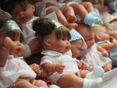 A collection of realistic baby dolls are arranged closely together. The dolls have various expressions and are dressed in different outfits, mostly in soft pastel colors. Some dolls have headbands, while others are in bonnets or knitted caps. The scene conveys a sense of innocence and childhood.