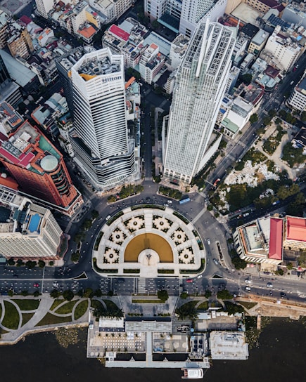 An aerial view of an urban area featuring tall skyscrapers and a large circular plaza with a prominent statue in the center. The cityscape is dense with numerous buildings, streets, and traffic visible. Green spaces and waterfront areas are also visible around the edges.