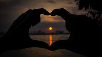 two hands making a heart shape with the sun setting in the background