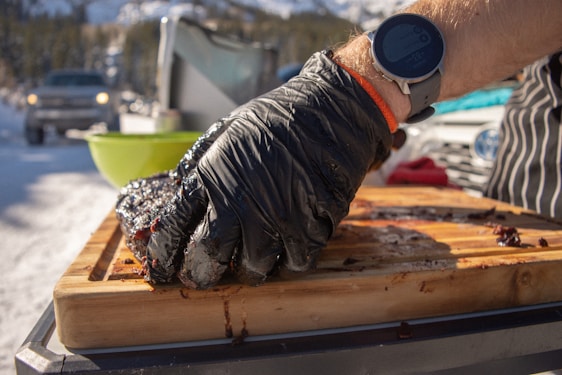 A person wearing a smartwatch and black gloves is handling a piece of cooked meat on a wooden cutting board. The setting appears to be outdoors in a snowy environment with vehicles and trees in the background.