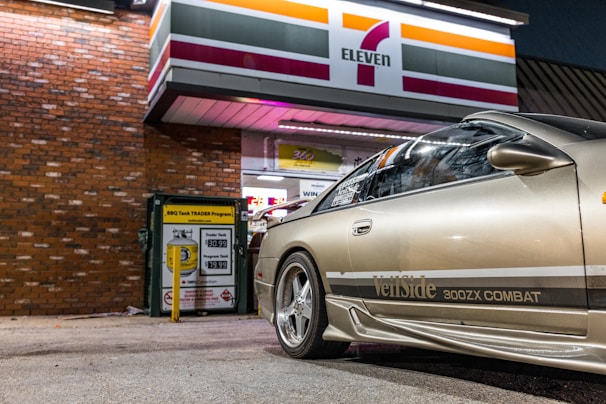 A silver sports car with VeilSide 300ZX COMBAT branding is parked in front of a brick wall under a 7-Eleven store sign. The scene is lit by artificial lighting, indicating a nighttime setting.