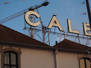 A large sign featuring the word 'CALE' is placed on top of a building with a tiled roof. A construction crane is visible in the background against a clear blue sky. Part of the building facade shows an arched window.