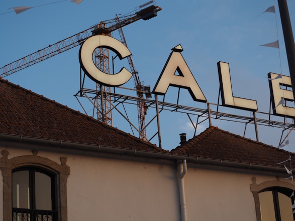 A large sign featuring the word 'CALE' is placed on top of a building with a tiled roof. A construction crane is visible in the background against a clear blue sky. Part of the building facade shows an arched window.