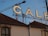 A large sign featuring the word 'CALE' is placed on top of a building with a tiled roof. A construction crane is visible in the background against a clear blue sky. Part of the building facade shows an arched window.