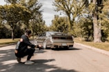 A photographer kneels on a suburban street capturing a sports car with a sleek design. The car is parked on the road surrounded by lush green trees and a clear sky above. The photographer is wearing a black hoodie with colorful design prints.