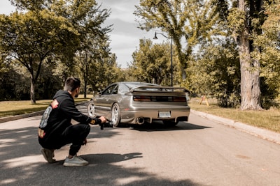A photographer kneels on a suburban street capturing a sports car with a sleek design. The car is parked on the road surrounded by lush green trees and a clear sky above. The photographer is wearing a black hoodie with colorful design prints.