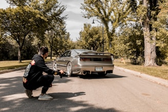 A photographer kneels on a suburban street capturing a sports car with a sleek design. The car is parked on the road surrounded by lush green trees and a clear sky above. The photographer is wearing a black hoodie with colorful design prints.