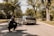A photographer kneels on a suburban street capturing a sports car with a sleek design. The car is parked on the road surrounded by lush green trees and a clear sky above. The photographer is wearing a black hoodie with colorful design prints.