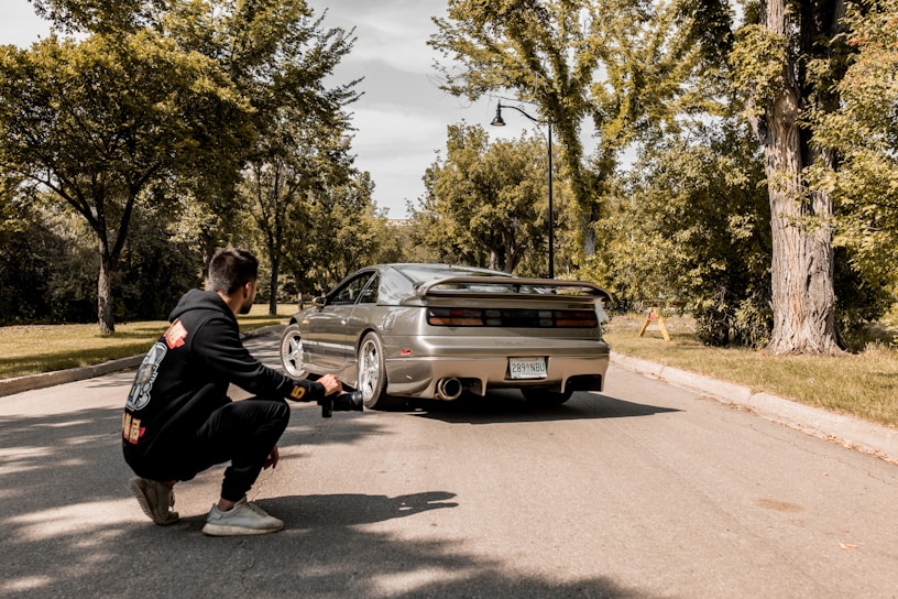 A photographer kneels on a suburban street capturing a sports car with a sleek design. The car is parked on the road surrounded by lush green trees and a clear sky above. The photographer is wearing a black hoodie with colorful design prints.