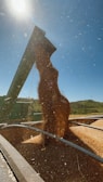 A stream of grain is being unloaded from a large agricultural machine into a vehicle or container. The scene takes place under a clear blue sky, with visible hills or fields in the background. Particles of grain create a dynamic motion as they fall.