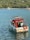 A small motorboat with a red canopy is floating on calm blue water. The boat is equipped with a life preserver and appears to be anchored. In the distance, there is a larger yacht near a tree-lined shore with dense green foliage.