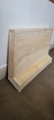 A wooden structure leaning against a white textured wall. The structure appears to be a handcrafted object, possibly a shelf or small storage unit, made from light-colored wood. The floor beneath is covered with a dark, textured material.