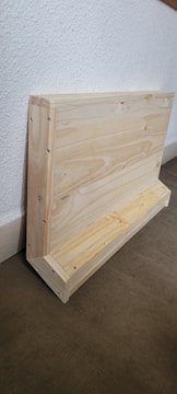 A wooden structure leaning against a white textured wall. The structure appears to be a handcrafted object, possibly a shelf or small storage unit, made from light-colored wood. The floor beneath is covered with a dark, textured material.