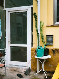 A small outdoor area features a white door with a glass panel, flanked by a tall cactus plant. Near the door are a round white table and a green watering can, along with a broom and a dustpan on the concrete floor. The yellow walls of the building include a mail slot and a window.
