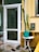 A small outdoor area features a white door with a glass panel, flanked by a tall cactus plant. Near the door are a round white table and a green watering can, along with a broom and a dustpan on the concrete floor. The yellow walls of the building include a mail slot and a window.