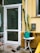 A small outdoor area features a white door with a glass panel, flanked by a tall cactus plant. Near the door are a round white table and a green watering can, along with a broom and a dustpan on the concrete floor. The yellow walls of the building include a mail slot and a window.