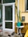 A small outdoor area features a white door with a glass panel, flanked by a tall cactus plant. Near the door are a round white table and a green watering can, along with a broom and a dustpan on the concrete floor. The yellow walls of the building include a mail slot and a window.
