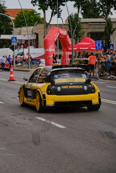 A bright yellow race car with the number 29 is speeding down a city street during a racing event. The car displays 'Visit Mangalia' on its rear. Spectators and officials are visible behind barriers, with red promotional banners and inflatable structures in the background indicating a sponsored event. The event is taking place on a well-maintained urban road lined with trees and buildings.