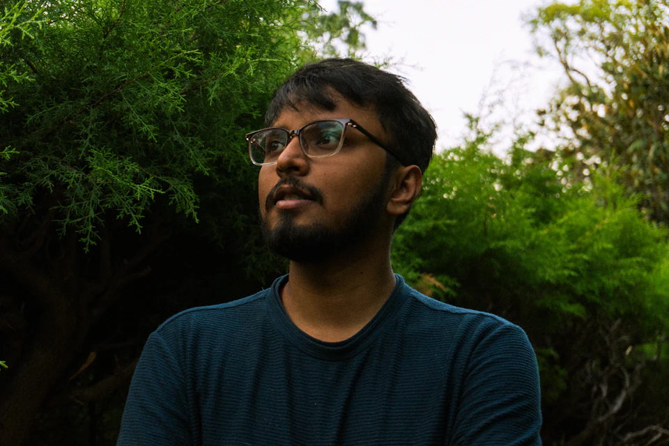 The image shows a man wearing glasses and a blue shirt, standing in front of a lush green bush with multiple branches. The man's gaze is directed upwards towards something beyond the frame of the photo.