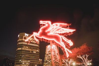 A bright red neon Pegasus sign is illuminated against the night sky. The sign is mounted on a metal structure with a cityscape visible in the background, including a tall building with office lights and a logo. Trees adorned with white string lights are visible at the lower right.