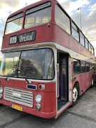 A vintage double-decker bus painted in a faded red color with white accents. The bus is labeled with the route number 025 and has 'Bristol' displayed on the sign above the windshield. The front features classic round headlights and a distinctive grille. The vehicle is parked and its door is open, showing a glimpse of the interior.