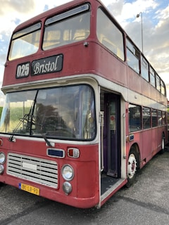 A vintage double-decker bus painted in a faded red color with white accents. The bus is labeled with the route number 025 and has 'Bristol' displayed on the sign above the windshield. The front features classic round headlights and a distinctive grille. The vehicle is parked and its door is open, showing a glimpse of the interior.
