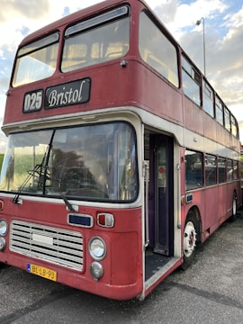 A vintage double-decker bus painted in a faded red color with white accents. The bus is labeled with the route number 025 and has 'Bristol' displayed on the sign above the windshield. The front features classic round headlights and a distinctive grille. The vehicle is parked and its door is open, showing a glimpse of the interior.