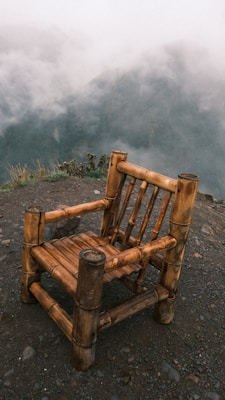 A rustic bamboo chair is placed on a gravel surface, positioned against a backdrop of misty mountains shrouded in clouds. The atmosphere is serene and natural, with the earthy tones of the chair contrasting against the soft, cloudy background.