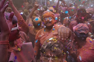 A lively and crowded celebration where people are covered in vibrant powder colors and confetti. Participants are energetically dancing or moving, with joyous expressions and gestures. Some individuals are wearing sunglasses and have their clothes stained with various hues.