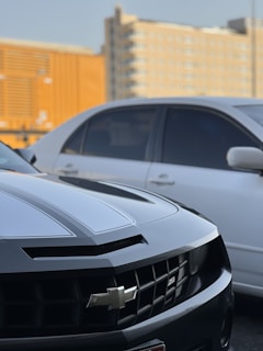 A close-up of the front section of a black Chevrolet car with a prominent grille and emblem. Another white car is parked beside it, and a high-rise building is visible in the background. The lighting suggests a late afternoon or early evening setting.