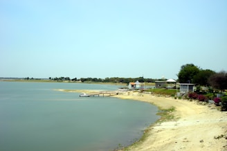 A calm lakeside scene with a small dock extending into the water. The shoreline is sandy with sparse vegetation and several trees. Buildings with colorful roofs are visible further inland, creating a small, serene park-like area.