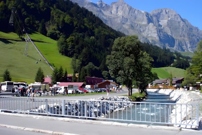 A picturesque mountain landscape with rugged peaks in the background, a lush green valley with trees, and a stream flowing alongside a road. There are people gathering near several buses and buildings situated at the base of the mountains. A bridge crosses the stream, and a ski lift structure is visible.