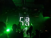 A dimly lit stage with a band performing. The background features a brick wall with 'rana' projected in large white letters. Green stage lights illuminate the scene, creating a vibrant atmosphere. Several people are visible watching the performance.
