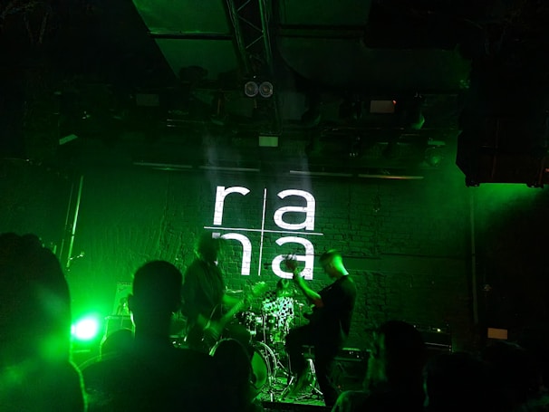 A dimly lit stage with a band performing. The background features a brick wall with 'rana' projected in large white letters. Green stage lights illuminate the scene, creating a vibrant atmosphere. Several people are visible watching the performance.