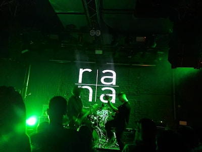 A dimly lit stage with a band performing. The background features a brick wall with 'rana' projected in large white letters. Green stage lights illuminate the scene, creating a vibrant atmosphere. Several people are visible watching the performance.