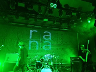 A dimly lit stage with musical instruments and performers in a green-hued setting. A brick wall serves as the backdrop featuring the word 'rana'. A drum set, microphone, and electric guitar are visible, with green lighting creating a moody atmosphere.