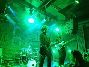 A live music performance features a drummer at the back with a checkered shirt playing drums, a guitarist with curly hair in the center, and a bassist in glasses on the right. Stage lights cast a vivid green hue over the scene, highlighting the performers and instruments. An audience member is visible in the foreground, capturing the performance on a smartphone. The background includes exposed brick walls and professional audio equipment.