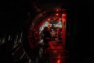 A dimly lit airplane cockpit with a red hue highlights the intricate controls and instruments. A person is seated in the pilot's chair, wearing a cap, in a focused posture facing the control panel. The atmosphere is both mysterious and intense, suggesting a late-night or early-morning setting.