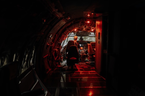 A dimly lit airplane cockpit with a red hue highlights the intricate controls and instruments. A person is seated in the pilot's chair, wearing a cap, in a focused posture facing the control panel. The atmosphere is both mysterious and intense, suggesting a late-night or early-morning setting.