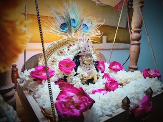 A decorative deity idol adorned with a crown and jewelry is placed in a wooden swing cradle filled with white and pink flowers. Peacock feathers are artistically positioned in the background, enhancing the vibrant and spiritual setup.